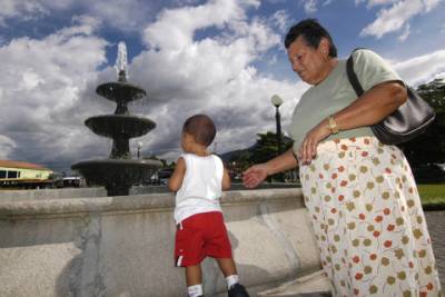 An elderly woman watches her grandson while he plays by a fountain in the center square in La Fortuna.